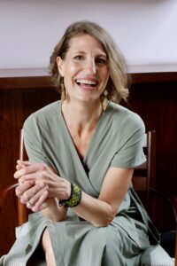 Shoshanna Grossman-Crist - white woman, blond, short hair leaning forward in a chair and smiling. 